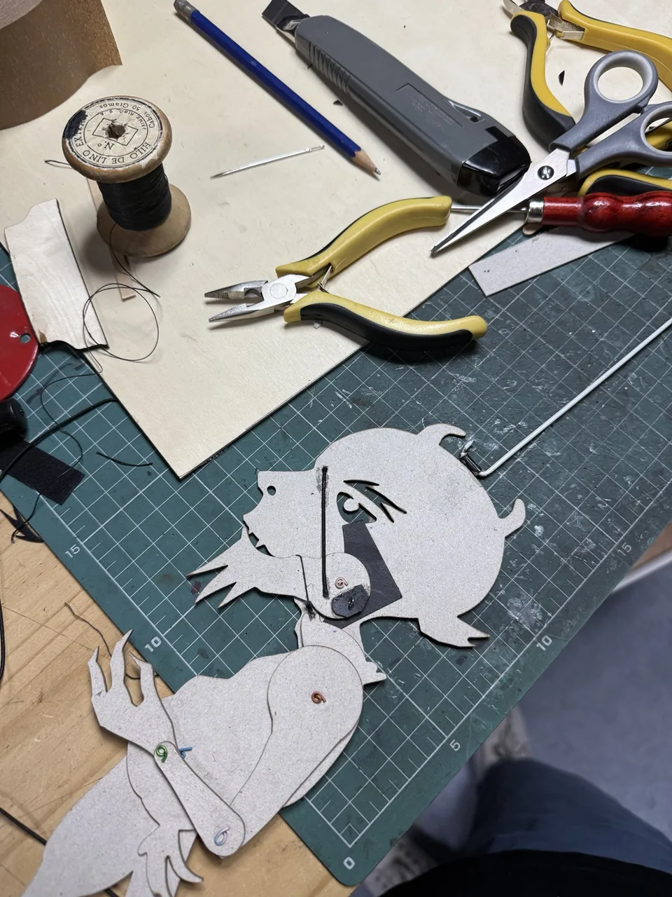 Silueta articulada de Cojuelo sobre la mesa del taller, junto a hilo, alicates y herramientas de construcción.